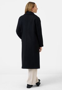Cappotto lungo nero in misto lana, con colletto rever, spalle scese e silhouette dritta, indossato sopra pantaloni beige e stivali neri chunky.