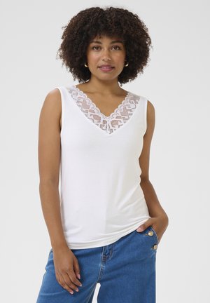 Mujer con cabello rizado, vestida con una blusa blanca sin mangas con detalle de encaje y jeans azules, de pie con una mano en el bolsillo contra un fondo blanco.