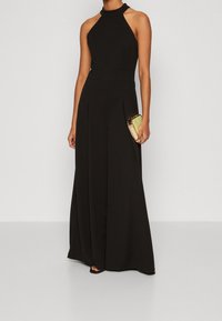 Mono negro con cuello halter y piernas anchas, tejido texturizado y pliegues a medida. Combinado con un pequeño clutch dorado.