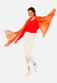 Un pull à col en V rouge associé à un pantalon crème. Une écharpe orange à bords effilochés tombe élégamment. Des baskets blanches complètent l'ensemble.