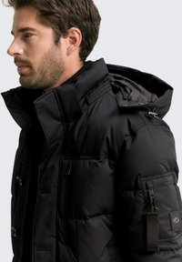 Černá bunda s péřovým vzhledem, vysokým límcem, velkými knoflíky a kapsami na zip. Nabízí texturovanou tkaninu a sportovní designové prvky.