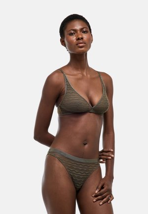 Femme portant un soutien-gorge triangle texturé vert olive et une culotte assortie, posant devant un fond clair uni, debout avec une main dans le dos.