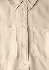 Camisa de color beige claro hecha de tejido suave, con un cuello puntiagudo, seis botones y dos bolsillos en el pecho con detalles de costura recta.