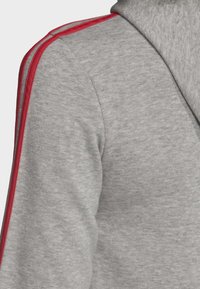 Felpa grigia con una texture morbida, caratterizzata da righe rosse lungo le spalle e il collo. Design con cappuccio, vestibilità casual.