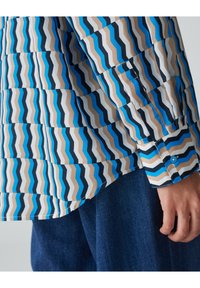 Kolorowa koszula z długim rękawem w zygzakowaty wzór w odcieniach niebieskiego, beżowego i czarnego, wykonana z gładkiego materiału, z mankietami na guziki i zaokrąglonym dołem.