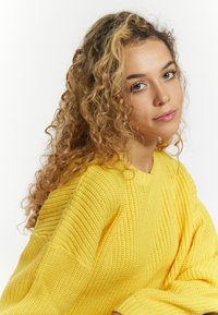 Strickpullover in leuchtendem Gelb, mit lockerer Passform, geripptem Textur und weiten Ärmeln. Nahaufnahme der Stoffdetails und des Kragens.