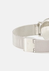 Silver mesh-armbandsur med metallspänne, som har en texturerad rem och släta detaljer på baksidan.