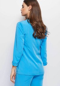 Lichtblauw op maat gemaakt blazer met een slank ontwerp, lange mouwen, een kraag en met knopen afsluitbare manchetten. Het materiaal lijkt glad en gestructureerd.