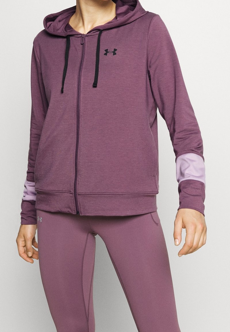 Personne portant un sweat à capuche violet zippé et des leggings assortis, avec des coudières violet clair et un logo noir sur la poitrine et la cuisse.