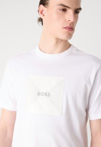 Balta medvilninė T-marškiniai su dideliu, tekstūruotu baltu kvadratu, turinčiu įstrižas linijas ir žodį "BOSS" centre. Trumpomis rankovėmis, apvaliu kaklu.