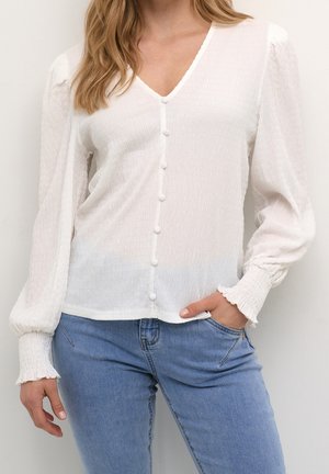 Weiße V-Ausschnitt-Bluse mit Struktur, überzogenen Knöpfen, aufgeblähten langen Ärmeln und gesmokten Manschetten, getragen mit blauen Jeans, Hand in der Tasche.