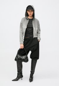 Šedá krátká mikina s kapucí s černým vzorovaným topem, volné černé kalhoty, černé kozačky po kolena a strukturovaná černá kabelka.