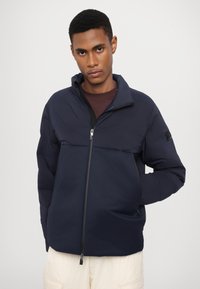 Jachetă puffer albastru marin cu guler înalt, fermoar complet și buzunare laterale. Textură netedă cu finisaj mat. Presentă un patch cu logo pe una dintre mâneci.