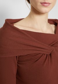 Top marrón cálido sin hombros hecho de tela suave, con un detalle anudado en el pecho y una textura lisa. Diseño simple y elegante.