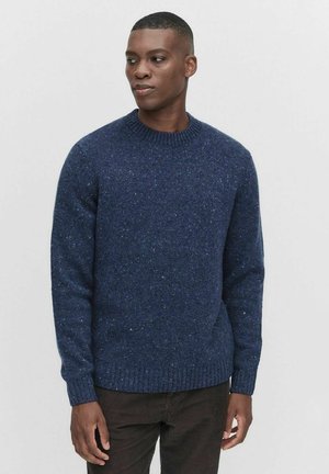 Marineblauwe gebreide trui met ronde hals, textured met spikkels, ribgebreide manchetten en zoom. Gedragen met donkerbruine broeken.