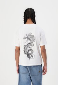 Hvit bomulls t-skjorte med et stort svart dragegrafikk på baksiden, med runde detaljer og japansk tekst integrert i designet.