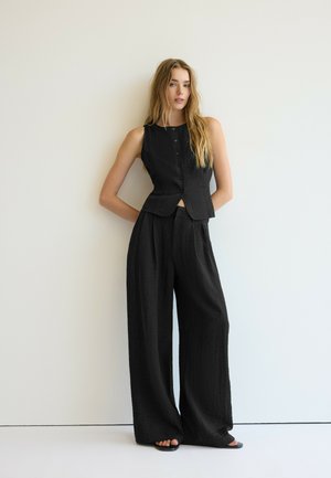 Femme aux longs cheveux blonds portant un gilet noir sans manches boutonné et un pantalon noir large, debout devant un mur blanc uni.