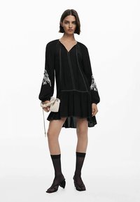 Juoda suknelė ilgomis rankovėmis, V formos iškirpte ir praskleistu apačiu. Yra balti siuvinėjimai ir sudėtingas siuvinėjimas ant rankovių. Papuošalai: maža rankinė.