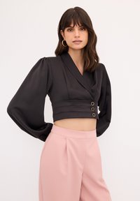 Schwarze kurze Bluse mit Puffärmeln, V-Ausschnitt und drei goldfarbenen Knöpfen in der Taille. Kombiniert mit hochtaillierten rosa Hosen.
