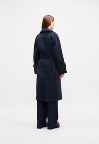 Cappotto di lana blu navy con cintura, dotato di un' unica fessura posteriore, ampi revers e tasche laterali. Indossato con pantaloni in denim scuro e scarpe nere spesse.