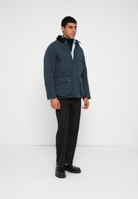 Men's navy quilted jacket with a zip and snap-button closure, two large front pockets, and a stand collar. Worn over a black turtleneck.