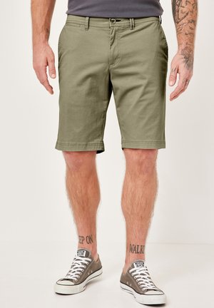 Khaki-Shorts aus Baumwolle, mit geradem Schnitt, seitlichen Taschen und umgeschlagenen Säumen. Getragen mit grauen Stoffschuhen.