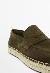 Olive green suede slip-on shoe with a woven jute sole, featuring a raised strap detail and contrasting stitching along the edge.