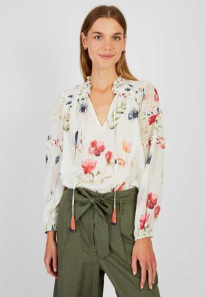 Frau trägt eine weiße Bluse mit Blumenmuster und Rüschen sowie olivgrüne, hoch taillierte Hosen, die mit einer Schleife in der Taille gebunden sind, und steht vor einem einfarbigen Hintergrund.