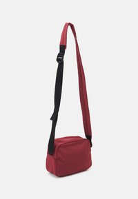 Borsa a spalla in tessuto rosso con esterno imbottito, forma rettangolare, tracolla nera regolabile e chiusura con zip sulla parte superiore. Texture liscia, design minimale.