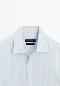Chemise bleu clair à boutons avec des rayures blanches verticales et un col à pointes écartées, soigneusement pliée pour mettre en valeur le col et les boutons.