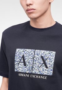 Άτομο που φοράει μαύρο t-shirt Armani Exchange με μεγάλο λογότυπο "A|X" σε μπλε και λευκό αφηρημένο σχέδιο στο στήθος.