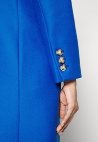 Cappotto di lana blu con una finitura testurizzata, dotato di un polsino sartoriale che mostra tre grandi bottoni rotondi e una silhouette slanciata.