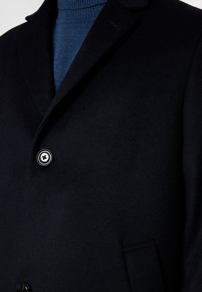 Dunkelblauer Mantel mit einem einzelnen Knopf, der über einen blauen gestrickten Rollkragenpullover geschlossen ist, sodass der Kragen und der obere Brustbereich sichtbar sind.