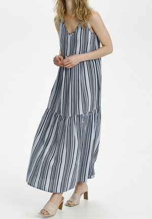 Femme portant une robe longue sans manches à rayures verticales bleues et noires, avec des sandales beiges à talons carrés ouvertes, debout devant un fond blanc.