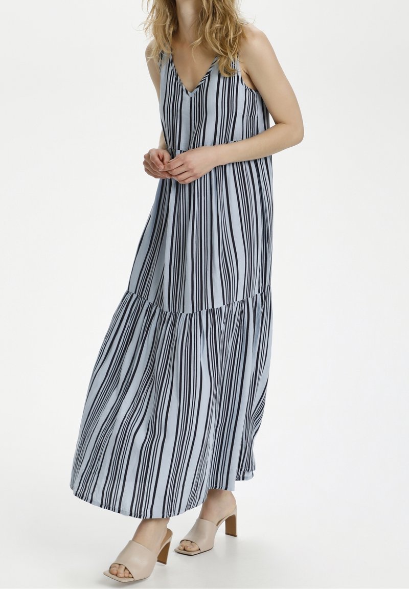 Femme portant une robe longue sans manches à rayures verticales bleues et noires, avec des sandales beiges à talons carrés ouvertes, debout devant un fond blanc.