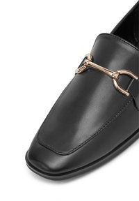 Čierne kožené loafery so štvorcovým špičkou, na ktorých je umiestnený zlatý kovový akcent na hornom diele. Hladká textura a elegantný dizajn.