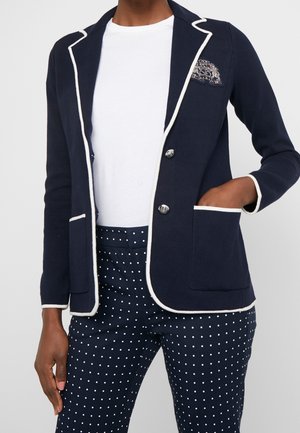 Person trägt marineblauen Blazer mit weißem Besatz und silbernen Knöpfen, weißes Hemd und marineblaue Hose mit weißen Punkten, Hand in der Blasentasche.