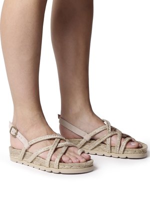 Two feet wearing beige braided strap sandals with buckle closure and textured sole, shown against a white background.