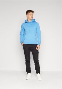 Felpa con cappuccio blu con tasca frontale, abbinata a pantaloni cargo neri con tasche laterali e sneakers bianche.