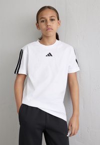 Weißes Baumwoll-T-Shirt mit kurzen Ärmeln, das mit dem schwarzen Adidas-Logo und drei schwarzen Streifen an den Schultern versehen ist, kombiniert mit schwarzen Jogginghosen.