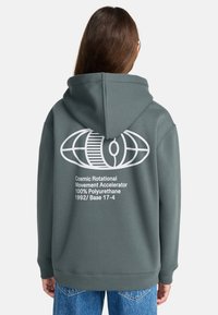 Person, die einen grünen Hoodie mit weißem geometrischen Design und dem Text "Cosmic Rotational Movement Accelerator 100% Polyurethane 1992/ Base 17-4" auf dem Rücken trägt.