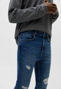 Tmavě modré džínové kalhoty s úzkým střihem, s trhanými detaily a klasickým designem s pěti kapsami, spárované s volným šedým tričkem s dlouhým rukávem.