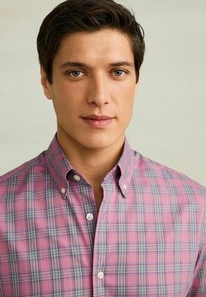 Junger Mann mit kurzen dunklen Haaren und blauen Augen, der ein rosa-grau kariertes Hemd mit Knöpfen vor hellem, einfarbigem Hintergrund trägt.