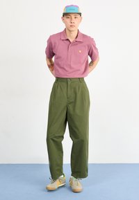 Cămașă polo roz deschis cu un logo mic, pantaloni verzi olive și adidași bej cu accente colorate. Material texturat, croială casual.