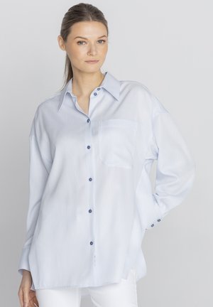 Młoda kobieta z włosami związanymi do tyłu, ubrana w jasnoniebieską, oversize koszulę zapinaną na guziki oraz białe spodnie, stojąca na jednolitym szarym tle.