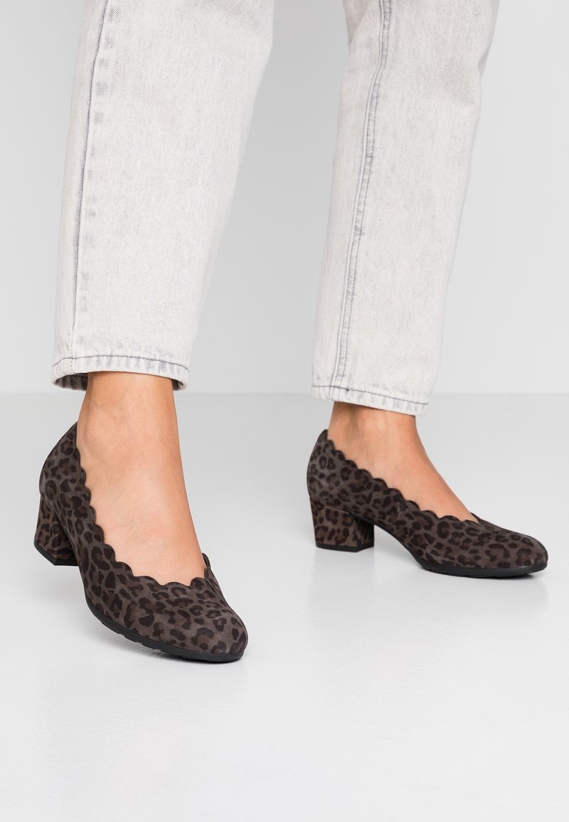 Femme portant un jean délavé clair et des chaussures à petit talon carré imprimé léopard brun foncé avec des bords festonnés sur un sol blanc.