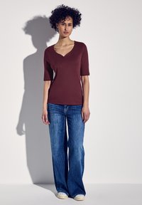 Bordowa bluzka z dekoltem w serek i krótkimi rękawami, zestawiona z szerokimi niebieskimi dżinsami. Modelka stoi na neutralnym tle.