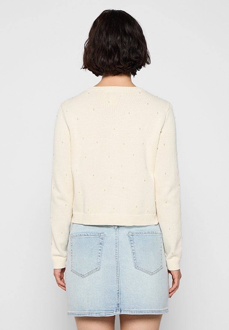 Cremfarbener Strickpullover mit kleinen Verzierungen, runder Ausschnitt und langen Ärmel, kombiniert mit einem hellblauen Jeansrock.