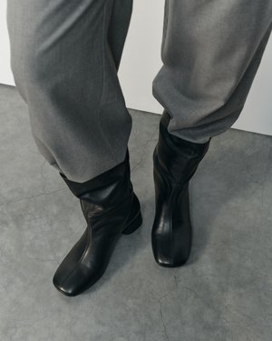 Personne portant un pantalon gris ample rentré dans des bottes en cuir noires mi-mollet, debout sur un sol en béton gris.