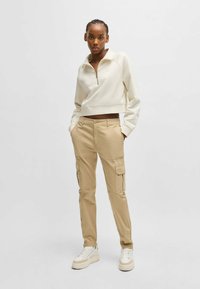 Felpa corta zip-up beige chiaro con colletto arrotondato, abbinata a pantaloni cargo khaki con tasche laterali e vestibilità slim. Sneakers bianche.